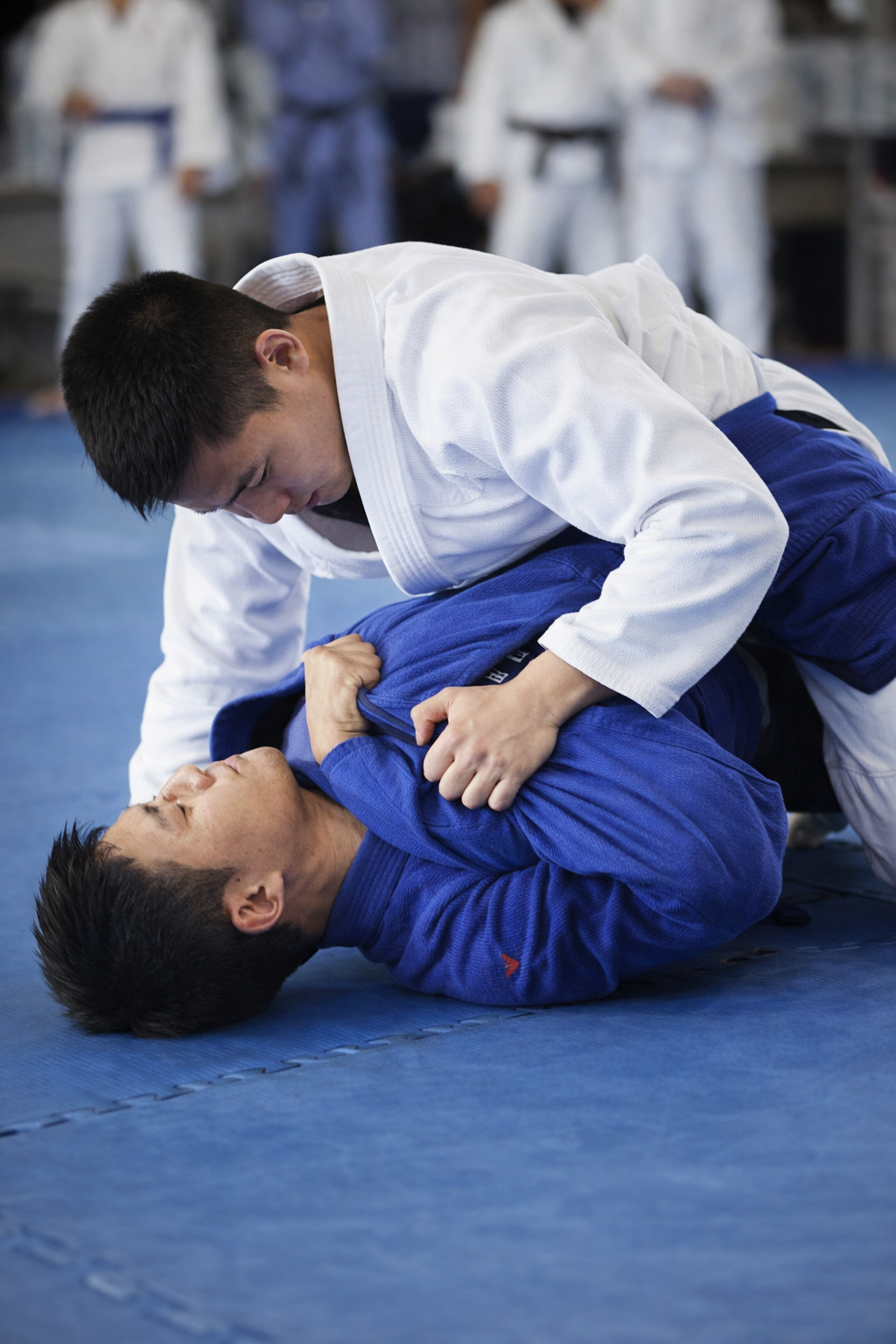 Two jiu jitsu practitioners sparring on a mat, with one in a white gi controlling an opponent in a blue gi during training.