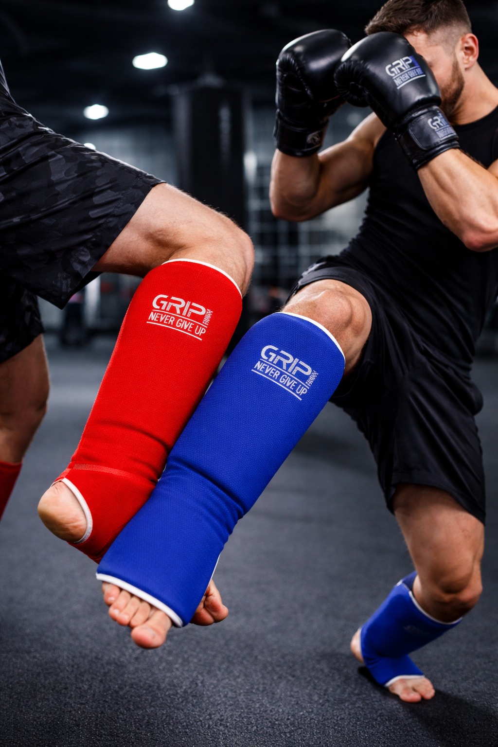 Two athletes sparring in a gym wearing red and blue shin guards, performing a kick and block with focused stance and blurred training background.