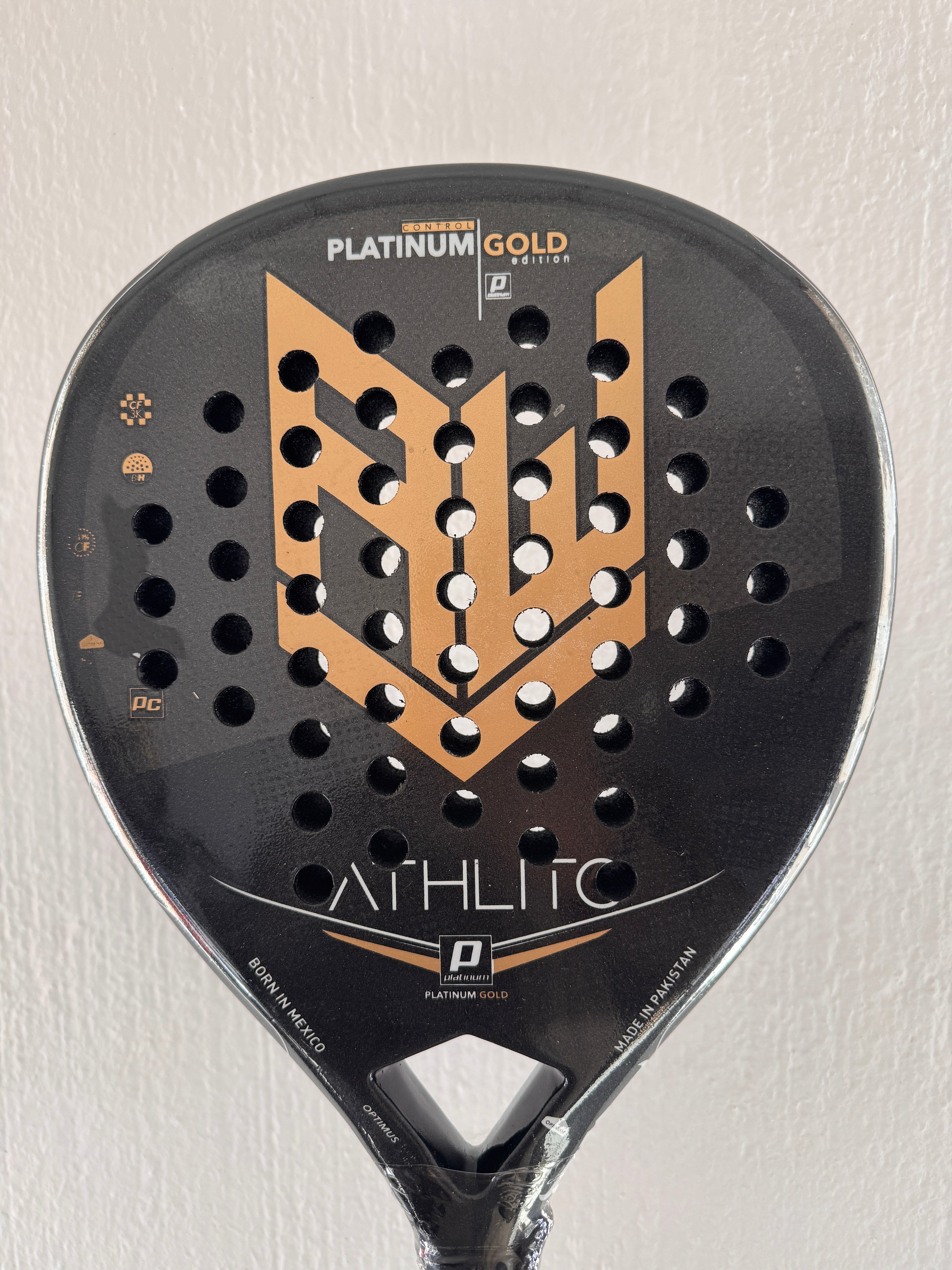 A close-up of a black and gold padel paddle face with the brand name 'ATHLITC' and model 'PLATINUM GOLD' printed on it, set against a plain off-white wall.