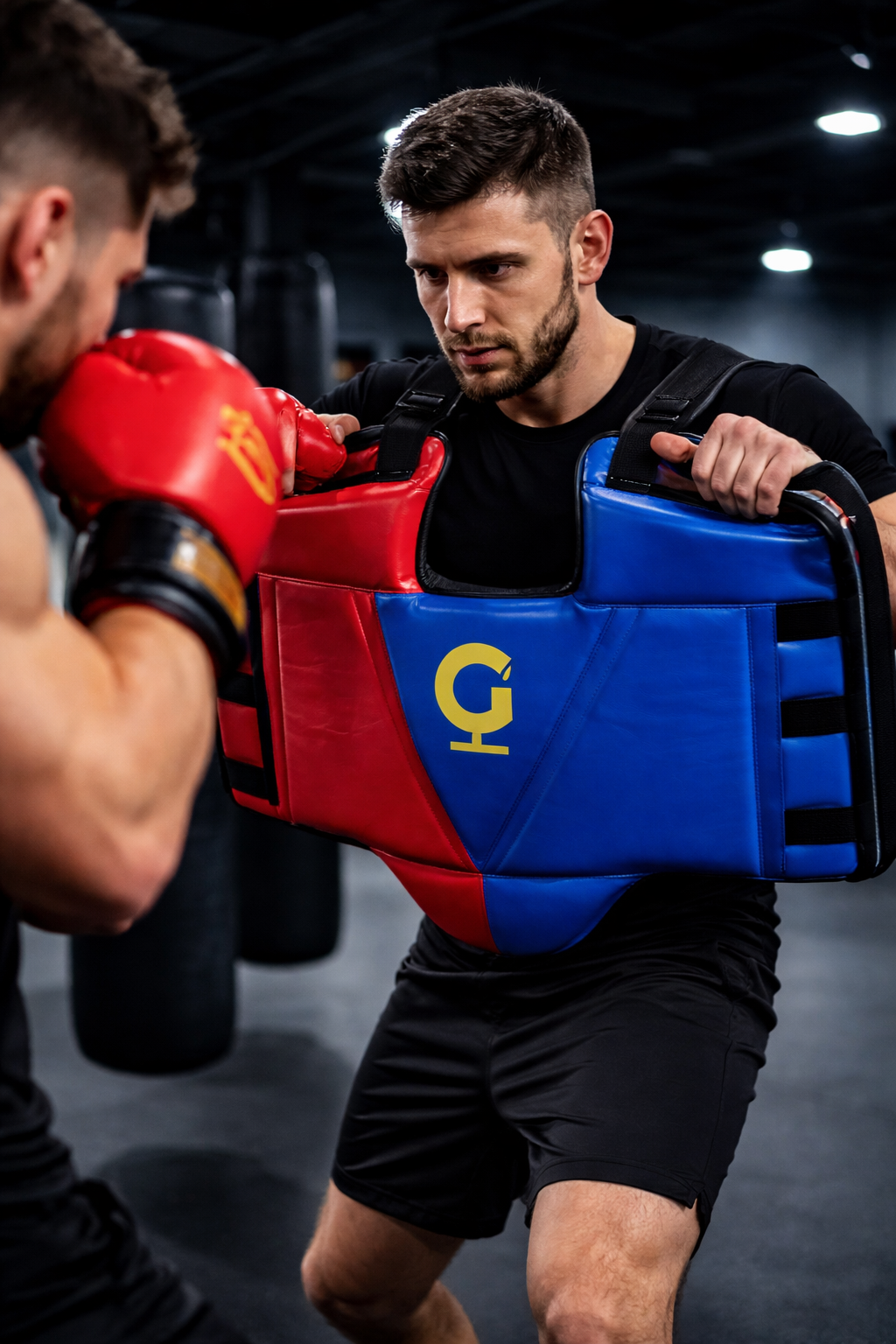 Boxing trainer wearing red and blue chest guard holds position while a boxer in gloves throws a punch during a gym training session.